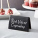 A black rectangular Acopa table tent sign with white text on a table in front of red velvet cupcakes.