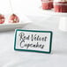 A teal ceramic table tent sign with a decal border on a table next to a red velvet cupcake.