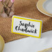 A yellow ceramic table tent sign with a border and a name tag on a table.