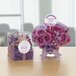 A bouquet of purple flowers in a glass vase on a table with a box of confetti with Avery 2 1/2" round scalloped labels on it.