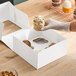 A gloved hand is placing a decorated cupcake into a white cardboard box with a four-slot insert on a wooden surface.
