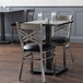 A Lancaster Table & Seating square wood table with a slate gray finish and two black chairs with black cushions.