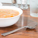A bowl of soup and a WNA Comet Reflections stainless steel look plastic soup spoon on a table.