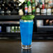 An Arcoroc stackable beverage glass filled with blue liquid with a lime and a black olive on the rim.