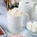 A close-up of a Tuxton white china c-handle mug with hot chocolate and marshmallows on a table.