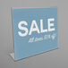 A Deflecto clear slanted sign holder displaying a blue sign with white text that says "Sale All Items 50% Off"