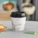 A Choice paper coffee cup with a bean print sleeve and lid with a cup of coffee and croissant on a table.