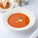 A CAC ivory china soup bowl filled with tomato soup on a table with a bread roll and a spoon.