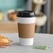 A white paper coffee cup with a brown band around it, a black lid, and a croissant on a table.
