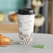 A Choice paper hot cup with a bean print sleeve and black lid on a table with a croissant.