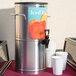 A Bloomfield stainless steel iced tea dispenser on a table with white cups.