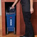 A woman in a black shirt and pants standing next to a blue Rubbermaid Slim Jim trash can with a black bag.