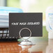 A Choice chrome menu / card holder with a round base holding a black sign that says "face mask required" on a table.