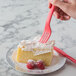 A hand holding an Eco-Products coral compostable plastic fork over a piece of cake with whipped cream and raspberries.