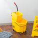 A Rubbermaid yellow mop bucket with a side press wringer and mop on a wood floor with a yellow caution sign.