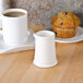 A white Tuxton creamer filled with milk on a table with a cup of coffee and a muffin.