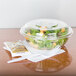 A salad in a Dart clear plastic bowl with a fork and spoon on a table.