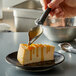 A hand using a Mercer Culinary precision spoon to pour caramel sauce over a piece of cheesecake.
