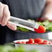 A person using Vollrath stainless steel tongs to hold a tomato over a bowl of tomatoes.