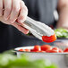 A person using Vollrath stainless steel scalloped utility tongs to hold a tomato.