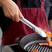 A hand uses Vollrath stainless steel utility tongs to hold a bacon strip over a skillet.