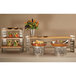 A buffet table with a Rosseto stainless steel riser stand holding meat and vegetables with flowers on the table.