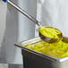 A person uses a Vollrath Blue Perforated Spoodle to scoop yellow food into a bowl.