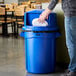 A person putting a plate into a blue Rubbermaid dome top trash can.