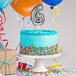 A white birthday cake with a silver number "6" on top and silver balloon decorations.