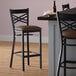 A Lancaster Table & Seating black metal cross back bar stool with a dark brown vinyl cushion.