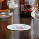A glass of beer on a 3 1/2" white round paper coaster on a table.