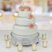 A Tabletop Classics nickel-plated cake stand holding a white wedding cake with pink flowers on a table.