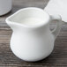 A Tuxton white porcelain creamer with a handle on a wood table filled with milk.