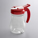A clear glass Vollrath teardrop syrup server with a red lid.