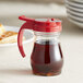 A clear glass Vollrath syrup server with a red lid and handle.