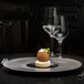 A RAK Porcelain gourmet flat plate in black and silver with food on it on a table with two wine glasses.