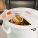 A hand pouring a plastic container of food into a white Rubbermaid ingredient bin.