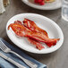 An Acopa narrow rim oval stoneware platter with bacon on a table with a fork and knife.