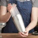 A woman in an apron using an Acopa stainless steel shaker to mix a cocktail.