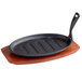 A black cast iron oval fajita skillet with a wooden gripper on a wooden tray.