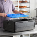 A black insulated food pan carrier with a blue ice board being placed inside, designed for keeping food hot or cold during transport.
