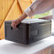 A person holding a black plastic CaterGator food pan carrier on a table.
