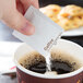 A hand holding a small white packet of sugar being poured into a cup of coffee.