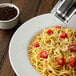 A bowl of spaghetti with tomatoes and whole black peppercorns on a table.