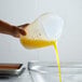 A hand using a Tablecraft 3-sided silicone measuring cup to pour yellow liquid into a bowl.