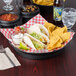 A table with a plate of tacos, chips, and a round deli server filled with red sauce.