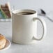 An ivory Acopa stoneware mug filled with brown liquid on a white saucer with a donut.
