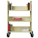 A Hirsh Industries putty book cart with 3 shelves full of books on wheels.