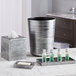 A silver cylindrical Focus Hospitality Parker wastebasket on a counter with small bottles of body care and a tissue box.