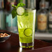 A Libbey stackable cooler glass filled with green liquid and a cucumber slice with a bowl of nuts on a table.